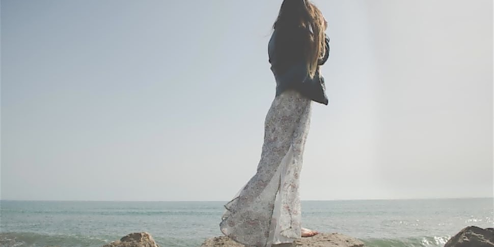 Frau in langem Sommerkleid steht auf einem Felsen und guckt Richtung Meer