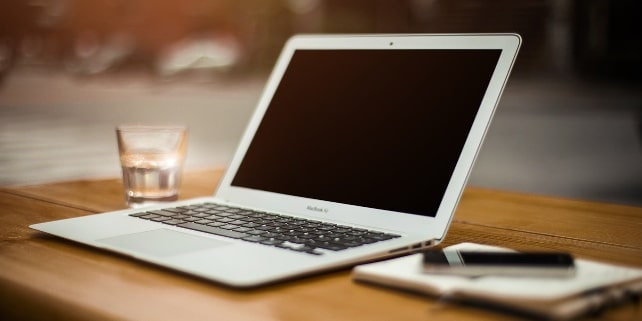 Ein MacBook steht zwischen einem Notizbuch und einem Glas Wasser Ein MacBook steht zwischen einem Notizbuch und einem Glas Wasser