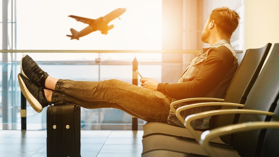 Mann am Flughafen schaut mit Beinen auf seinem Koffer aus dem Fenster zum Flugzeug