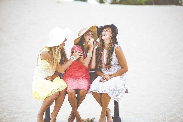 Drei Frauen mit Hüten und bunten Sommerkleidern auf Sitzbank am Strand.